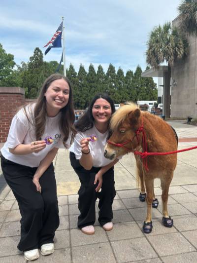 Two University of South Carolina students smiling beside a miniature therapy horse wearing shoes outdoors during the AutismConnect event