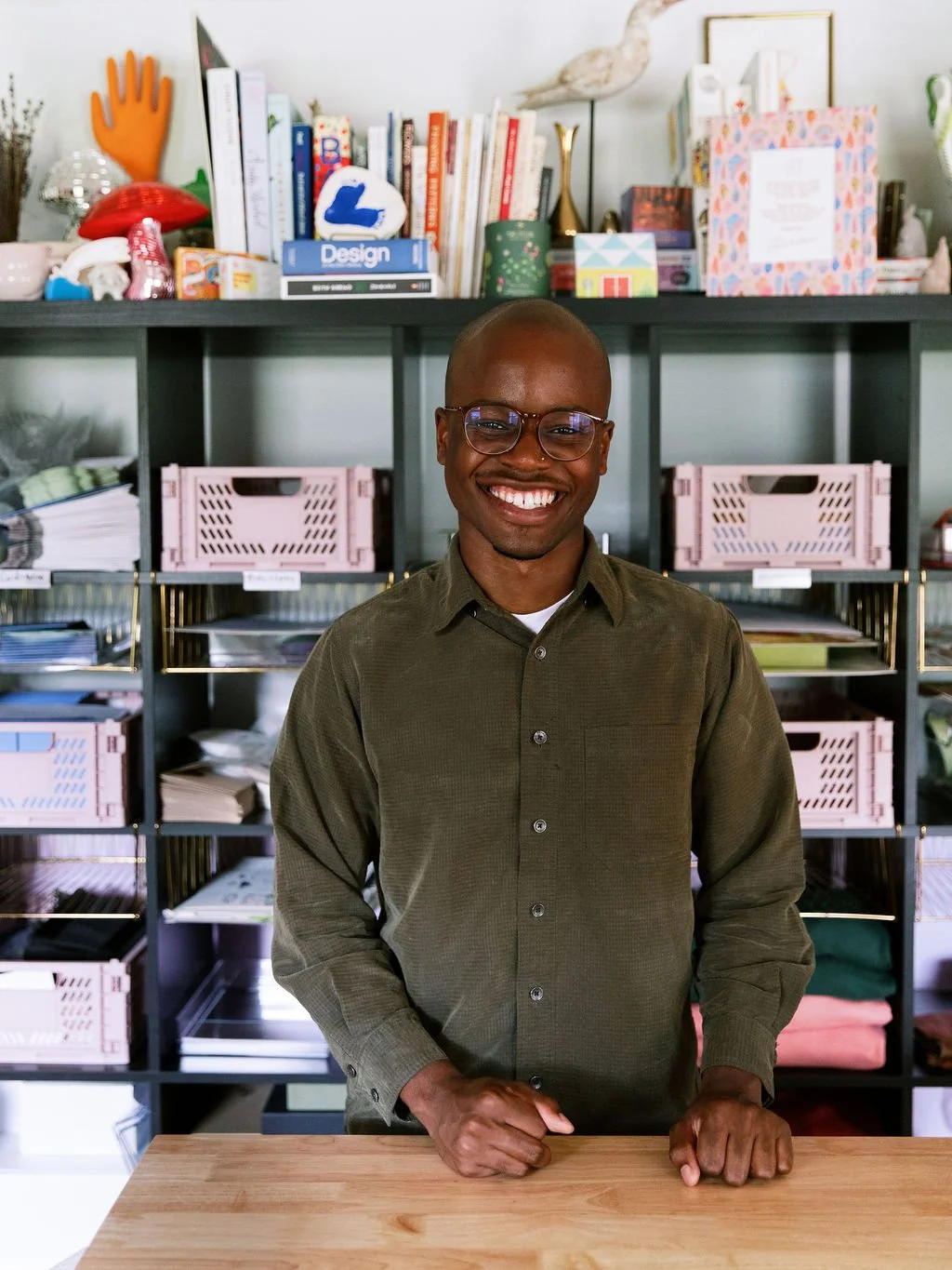 Smiling student wearing glasses and a green button-up shirt standing indoors in front of shelves filled with neatly arranged books or printed materials.