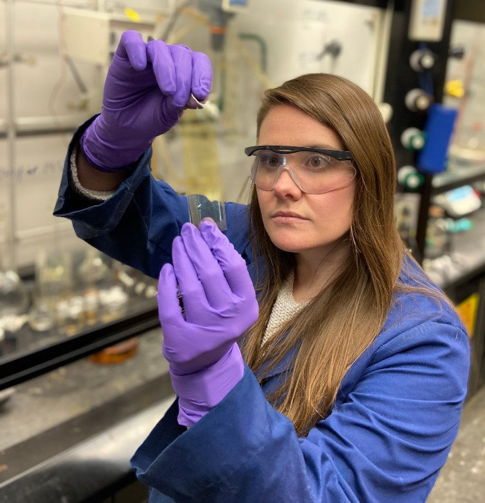 Wearing blue lab gloves and safety glasses, Laura Murdock holds up a thin piece of plastic film