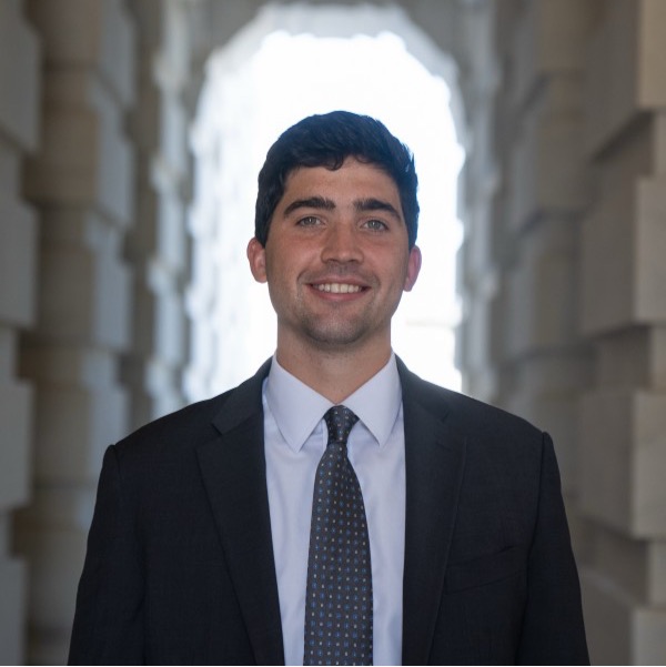 Portrait of a smiling man in a dark suit and tie standing beneath a stone archway.