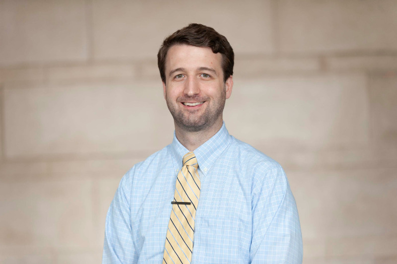 Matt McMillan wears a blue button up shirt and yellow tie and smiles at the camera