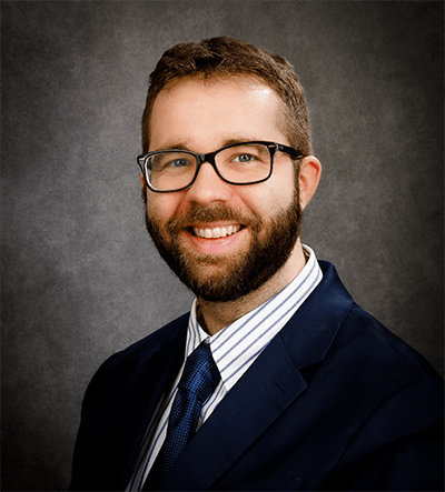 Portrait of Matthew Facciani, smiling and wearing glasses, a dark suit jacket, a striped dress shirt, and a navy blue tie. The background is a neutral gray studio backdrop.