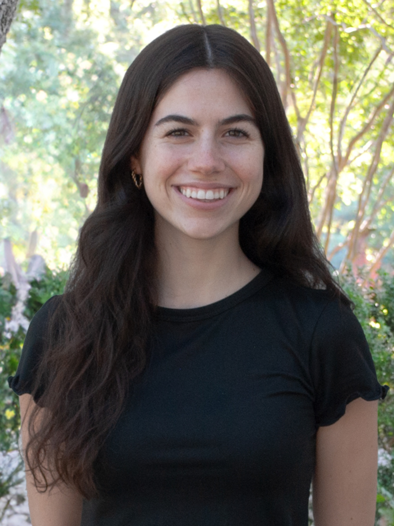 A young woman with long, wavy dark brown hair smiles warmly while standing outdoors. She is wearing a black short-sleeved top, and the background shows sunlit trees and greenery.