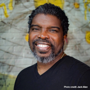 Darion McCloud is an African American man with short, curly black hair and salt and pepper facial hair. He wears a black shirt and smiles at the camera from in front of a white and yellow background. White photo text reads "photo credit to Jack Allen".