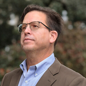 Ed Madden is a white man with short brown hair and black-framed glasses. He wears a light blue collared shirt and a brown suit jacket and is looking off-camera with a slight smile.
