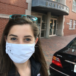 Juliana has long dark hair and wears a monogramed jacket and a mask, and she stands in front of the Boston Ballet building.