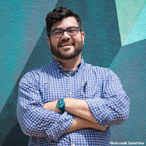 Lee Snelgrove is a white man with short, dark hair and dark facial hair and is wearing dark-rimmed glasses. He wears a blue and white plaid button-down shirt and smiles at the camera with an abstract wall painting behind him. White photo text reads "photo credit to Daniel Hare".