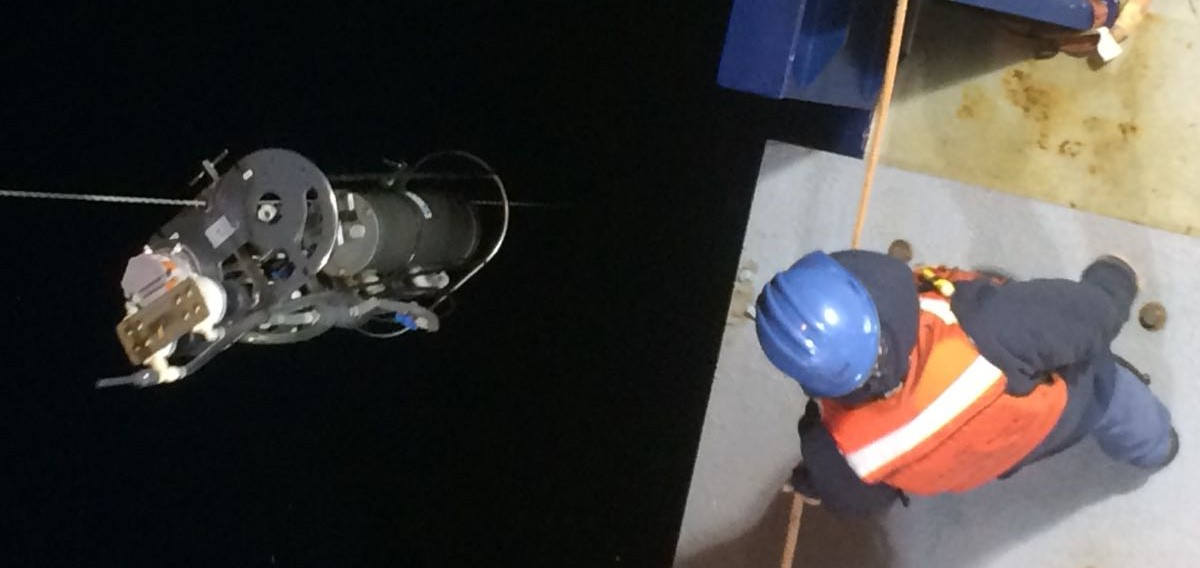 An overhead view looking down on the deck of a research cruise ship. A piece of research equipment is being lowered into the water as a researcher looks on.