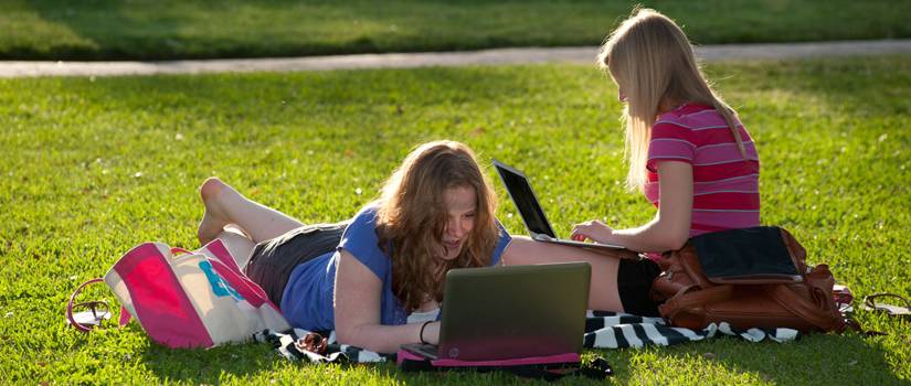 Two girls with laptops lounging on grass in the sun