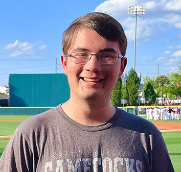 Zach McClure smiles while standing in a baseball stadium, wearing a University of South Carolina Gamecocks T-shirt. The field, players, and bright blue sky are visible in the background.