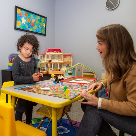 A clinician sits at a low table with a child client and observes the child playing with toys and board games.
