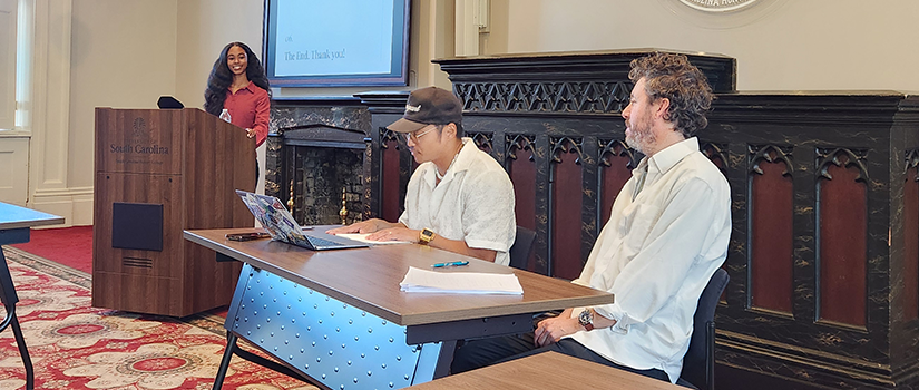 Religious Studies student Kamryn Carter presents her Honors thesis at a podium in a classroom, while two faculty members sit at a front table listening. A screen behind her displays a presentation slide that reads “The End. Thanks, you!” and the South Carolina Honors College emblem is visible on the wall.
