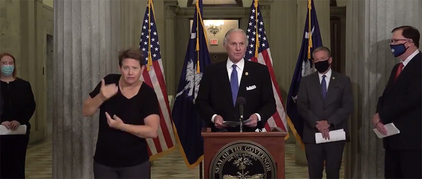 Henry McMaster holding a press conference.He is at the podium (without a mask). An interpreter is signing (without a mask). Other officials stand around (wearing masks).