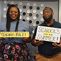 two African American teachers, one female, one male. One holds a sign that says "Teachers Rule!" the other a sign that says "School's Out for Summer!"
