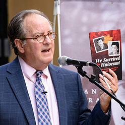 Frank Baker holding a copy of "We Survived the Holocaust" while talking into a microphone