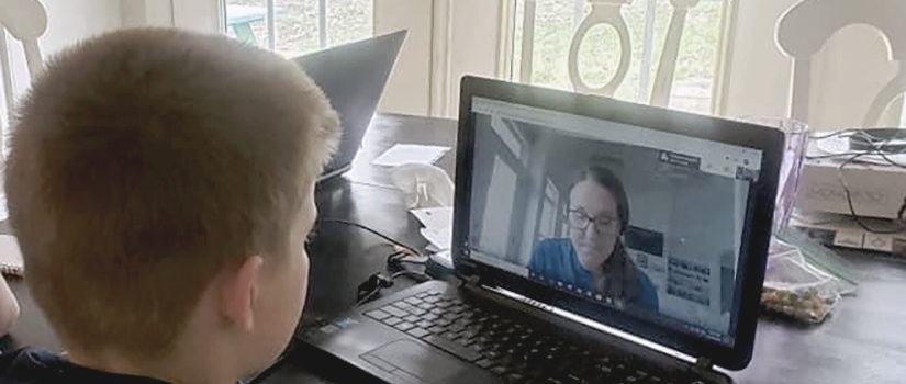 a child seated at a kitchen table observes a virtual lesson from a teacher on a laptop screen