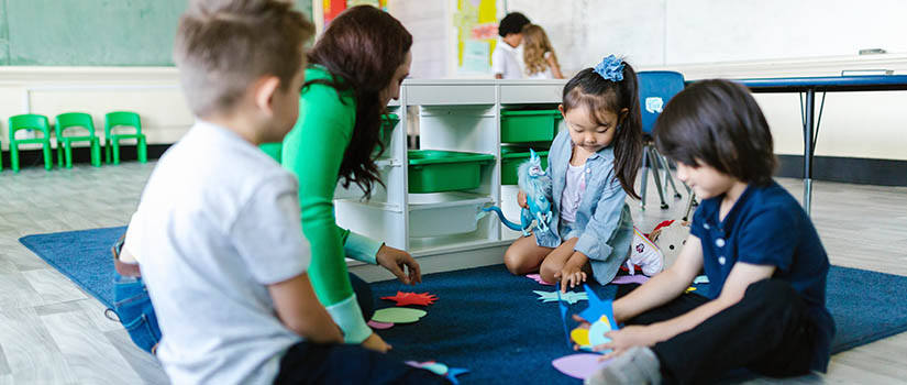 Teacher dressed in green leading a small group of very young children in an activity on the floor of a white classroom