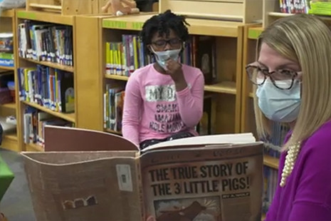 masked teacher reading book to maksed student