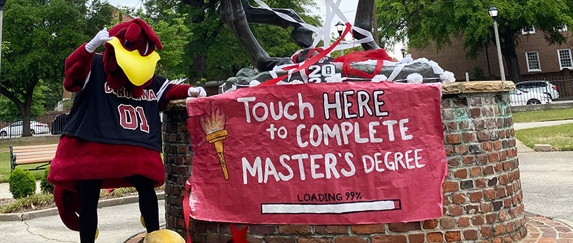 UofSc mascot Cocky pointing to a sign that says 'Touch Here to Complete Master's Degree' at the base of the Torchbearer statue.