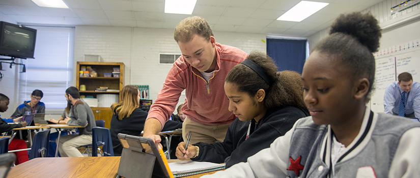 A teacher points to a tablet that a student is working on while another student smiles in the foreground.
