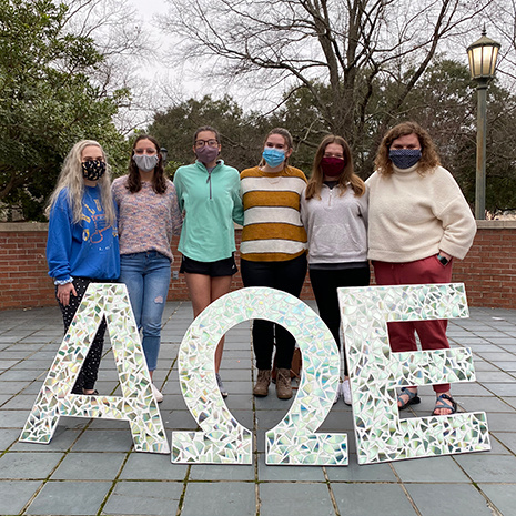 six women stand wearing face coverings behind large AOE letters