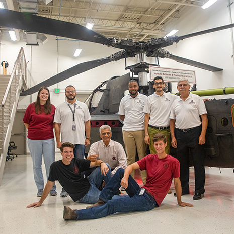 a group of faculty and students pose in front of the Apache helicopter in the McNair building