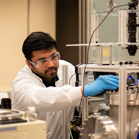 Sobhan wears a lab coat and safety glasses while working with equipment in the lab