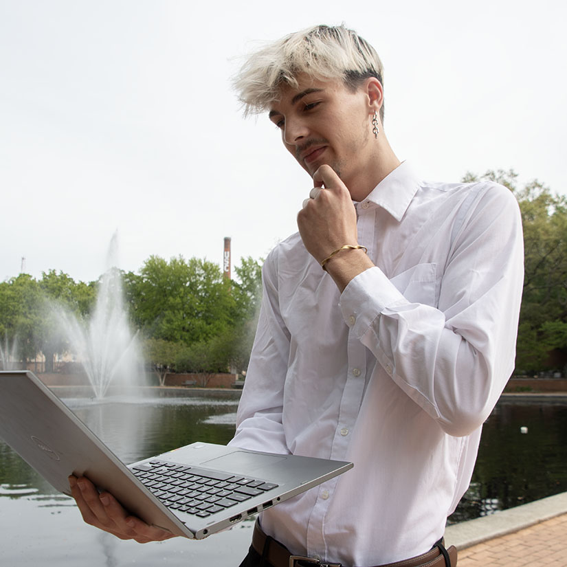 Student Rye Stahle-Smith looking at a laptop in front of the fountains next to the Thomas Cooper Library.