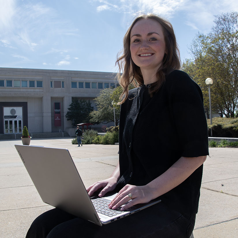 Sara Clark with a laptop with the entrance to the Swearingen Engineering Center in the background. 