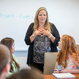 Female professor in a classroom in front of a whiteboard talking to students at a table in front of her.