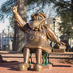 Bronze status of Cocky, the Gamecocks' bird mascot sitting on a bench with one hand on a pile of books and one hand in the air.