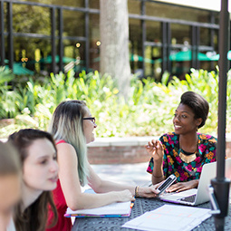 Two female students talking over paper and a laptop at an outdoor table surrounded by other students and greenery.