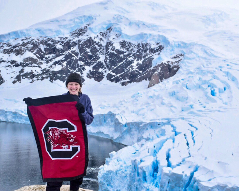 Caroline Cates holding a Gamecock flag in front of lake and icy mountain in Antarctica.