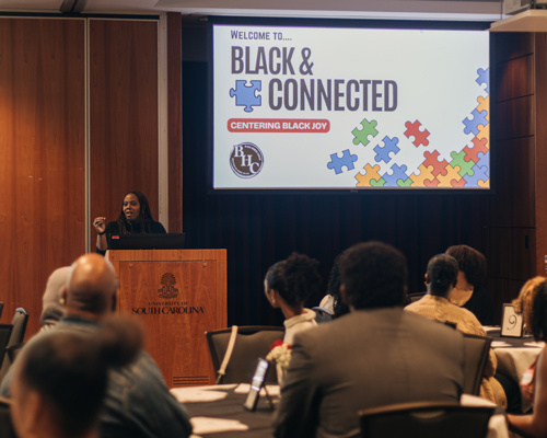 Black woman speaking to crowd behind a podium with a screen behind her that reads: Black & Connected: Centering Black Joy.