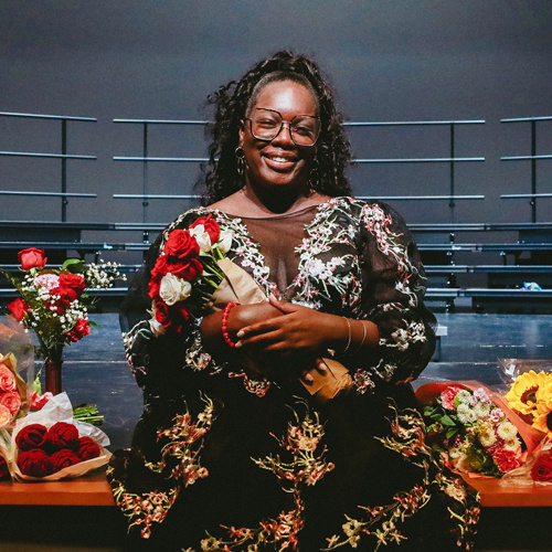 Smiling Black woman holding flowers with an empty stage behind her.