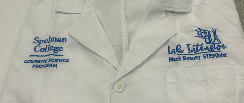 White lab coat with "Spelman College, Cosmetic Science Program," embroidered on the right side and "Lab Intensive, Black Beauty STEMinist" on the left side.