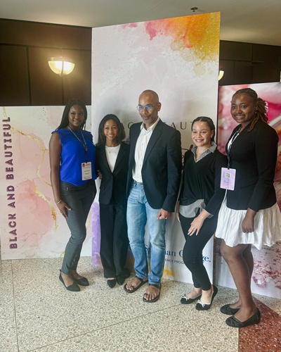 Kylie Mudie with a group of people posing for a photo. The pink and yellow watercolor photo backdrop reads, "Black and Beautiful," to the left side.