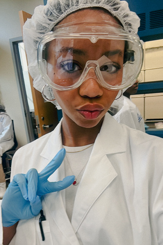 Selfie of Kylie Mudie in a white lab coat and protective lab gear as she poses her blue, gloved hands in a peace sign.