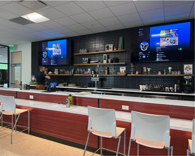 Wide view of man in a well-lit beverage station with two mounted TVs displaying the music playlist for the class.