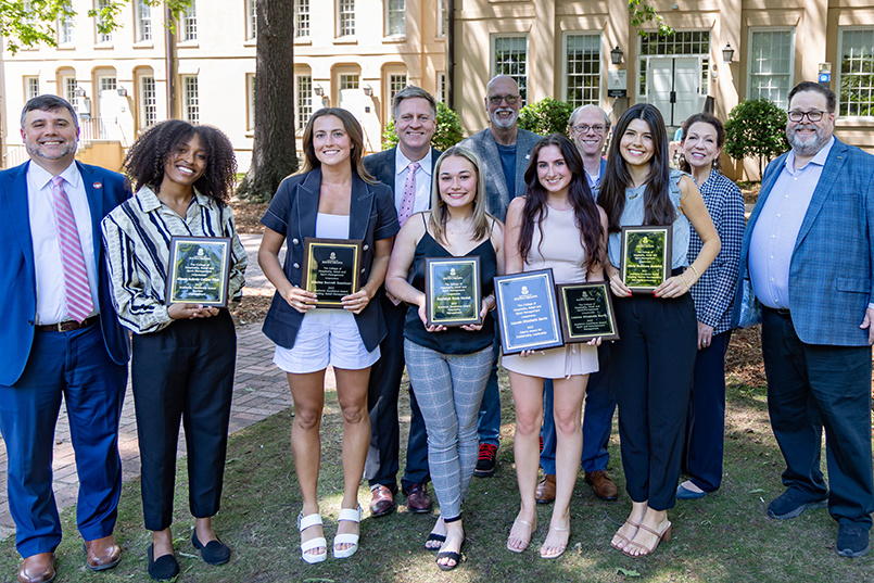 A group of faculty and students gather for a photo with students displaying their awards while attending a ceremony on the Horseshoe.