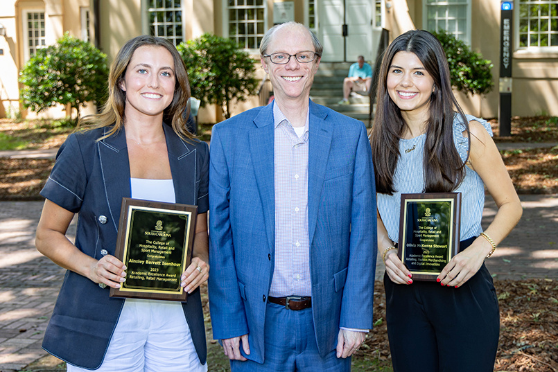 Two students show off their academic awards while posing for a photo with a faculty member.