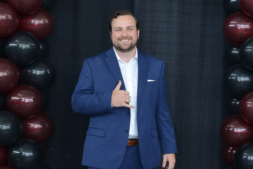 Joe Sasso throws up the Spurs Up gesture while standing in front of a black backdrop with garnet and black balloons on the sides.