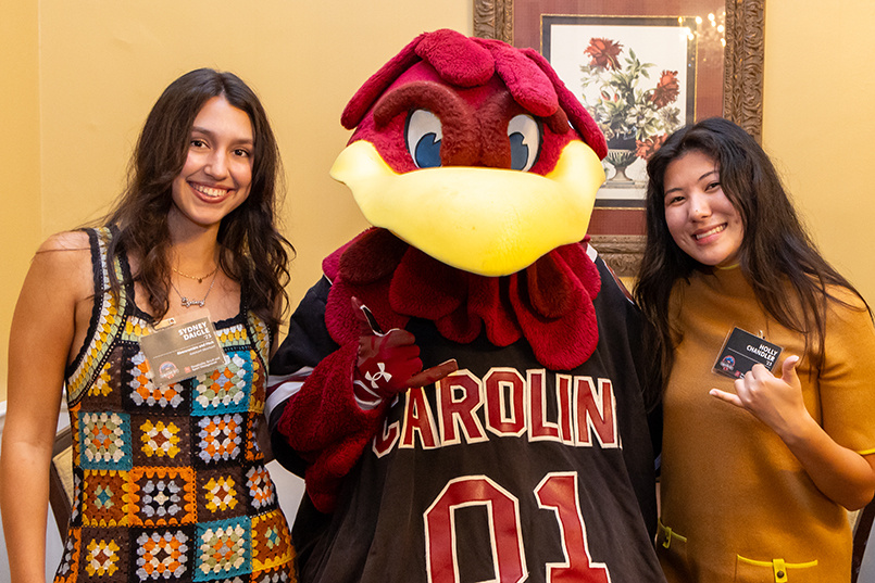 Two HRSM alums pose for a photo with USC mascot Cocky inside McCutchen House.