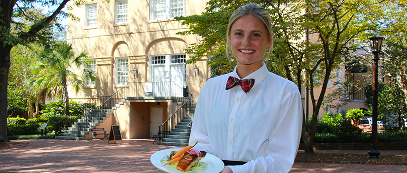 A female hospitality management student stands on the Horseshoe in front of the historic McCutchen House. She holds a delicious-looking plated meal.