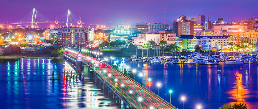 Charleston, South Carolina, illuminated at night with a view of the Cooper River bridge in the backgound and colored city lights reflecting on the river and marsh in the foreground.