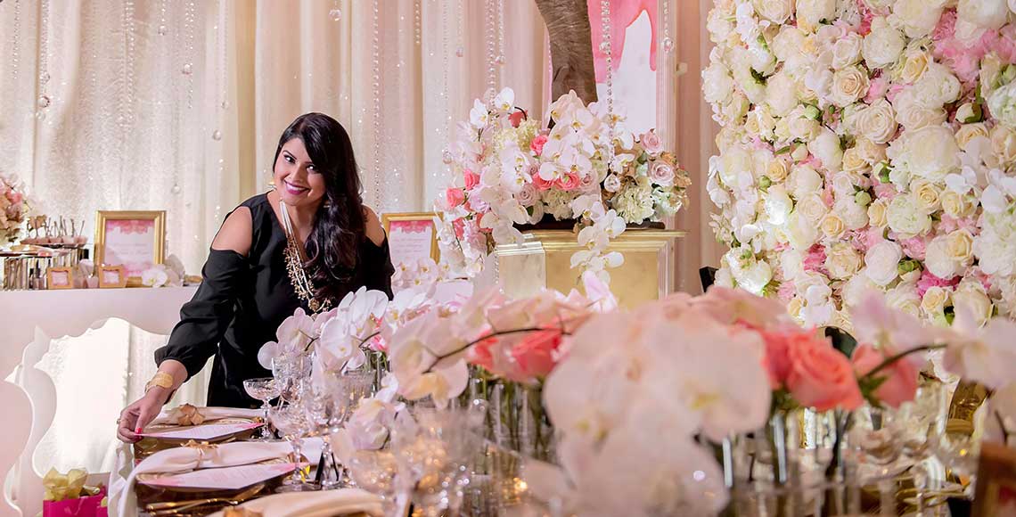Nirjary Desai, a hospitality management alumnus, stands in a wedding banquet room surrounded by roses, decorated trees and branches, and a sumptuous dining table