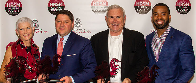 2019 Alumni Award winners stand in front of a step and repeat screen with the HRSM Alumni Society logo. Winners are Distinguished Service Award Winner Glenna Gillentine; Distinguished Alumni Award Winner Tom Miguel DeLozier; Distinguished Friend of the College Award Winner JDA; and HRSM Distinguished Young Alumni Award Winner Brandon Wade Ruth.
