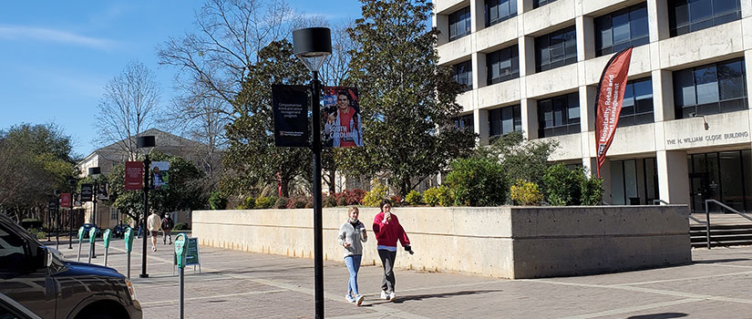 The Close-Hipp Building on a beautiful clear day with blue skies. two female students are walking with coffee chatting. HRSM brag banners of HRSM students hang in front of the building from the light posts, and welcome feather flag banners are displayed on the ground.