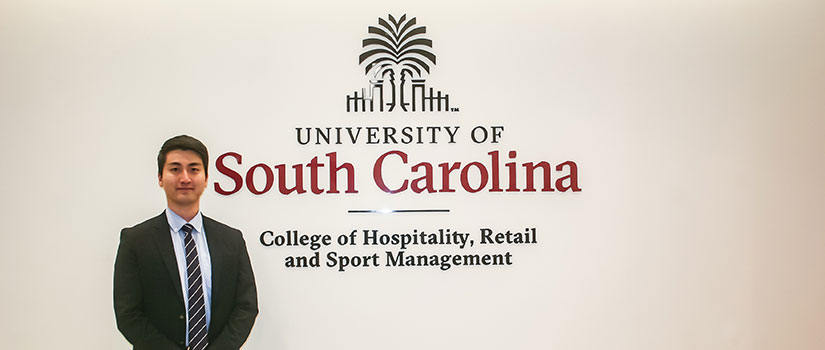 Ph.D. student in hospitality management, Henry Kim, stands beside the university's formal logo on the wall of the HRSM visitor center.