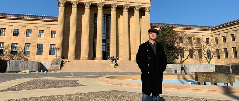 Guanlin Wu, a Beijing native, stands in front of the Philadelphia Museum of Art, a Greco-Roman classically designed building, on a sunny blue-sky day.